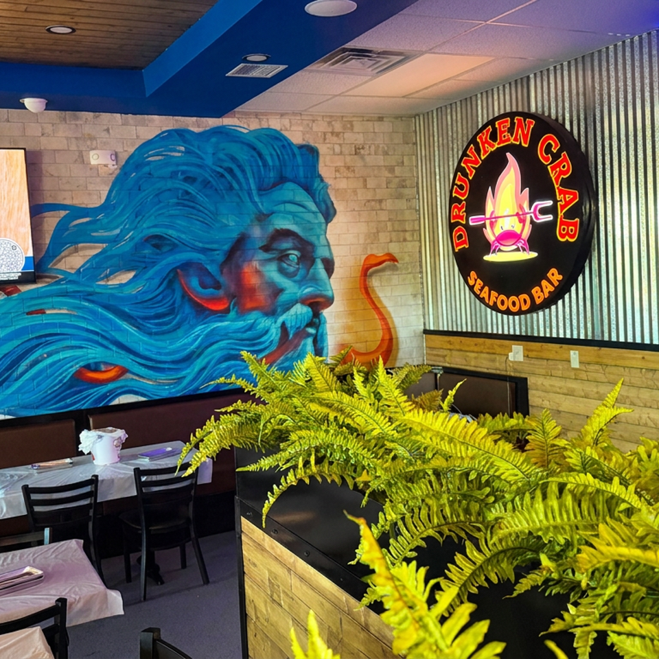 Colorful seafood restaurant interior featuring a large blue sea-god mural, Drunken Crab Seafood Bar sign, dining tables, and lush ferns.